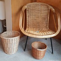 Wicker tub chair with two wicker waste baskets placed on carpet against a wooden wall
