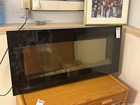 Full view of black electric wall-mounted fireplace with glass front sitting on wooden chest, plug visible.