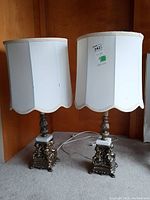 Pair of white scalloped fabric shade table lamps with ornate metal cherub figure bases against a wood paneled wall.