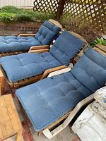 Photo of three wooden deck loungers with blue tufted cushions arranged outside on a brick patio near garden lattice and plants.