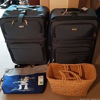 Two black Tracker soft-shell suitcases standing upright beside a blue and grey Canada themed tote bag with attached feather charm, and a woven basket tote on floor