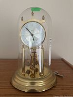 Front view of Kundo mantel clock with glass dome, showing clock face and pendulum beneath, placed on a wooden surface with winding key on the right.