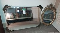 Two vintage wall mirrors by Peacock Mirrors shown standing against wall. One is large rectangular with decorative metal corners, other is oval with ornate gold carved frame.