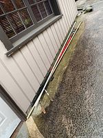 Three long pole pruners/trimmers leaning against exterior siding wall on pavement, showing poles and handles.
