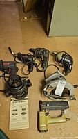 Set of six power tools displayed on floor: two drills, circular saw, router, pneumatic stapler, and instruction manual