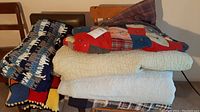 Side view of multiple folded quilts and crocheted blankets stacked on chairs showing various colors and patterns, including solid cream and patterned patchwork.