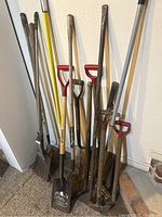 Full view of various yard tools including shovels with wooden handles and metal blades, rakes with metal tines, and edgers with red D-shaped handles, all showing weathered use.