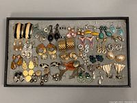 Full view of assorted pairs of costume earrings arranged in box tray, showing variety of styles and colors.
