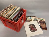 Red plastic milk crate filled to capacity with more than 60 vintage vinyl record albums, showing varied cover artwork including Christmas and pop music albums outside the crate.