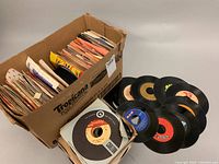 Photo of cardboard box filled with over 70 vintage 45 RPM vinyl records, some records placed outside the box showing various label colors and designs.