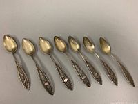 Photo showing top view of seven antique silver spoons arranged in row, highlighting the bowl shape and filigree handles with minor tarnish and wear.