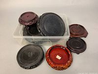 Eight vintage Chinese wooden planter bases displayed inside and outside a clear plastic bin showing different sizes, wood colors, and styles with carved details and legs on some pieces.