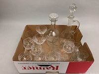 Assortment of crystal stemware including two lidded decanters, balloon wine goblets, votive glasses, measuring glass, and pitcher in a cardboard box.