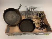 Photo showing entire lot including two cast iron skillets, three wrought iron candleholders, and a foldable metal toast rack in a cardboard box.