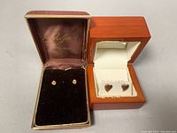 Two pairs of earrings displayed in their respective boxes: one in a brown velvet box with floral studs, the other in a wooden box with white lining holding the rose gold heart studs.