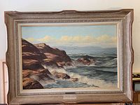 Full view of framed oil painting showing coastal cliffs, waves, sky with clouds, and birds flying. Frame mostly gold/beige with minor wear.