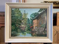Framed landscape oil painting displayed frontally showing building, pond, and forest scene