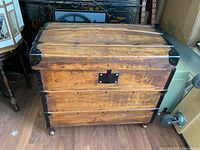 Front view of vintage wooden travel trunk showing wood slats, black metal corner reinforcements, latch area, and casters underneath.