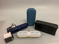 Five assorted portable Bluetooth speakers on a flat surface: Skullcandy Air Raid in dark blue, Beats Pill in white and gold, Borne BTSPK23 fabric speaker, a black rectangular speaker, and a small white/red cube speaker