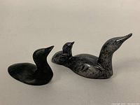 Two soapstone carved bird sculptures, one black and one mottled gray with a chick on its back, shown side by side on a flat surface.