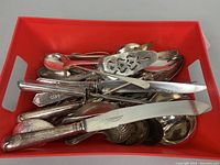 Assorted silver-tone vintage serving utensils including cake cutters, ladle, berry spoon, bread knife, and tongs in red bin