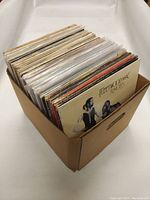 Vinyl records stored vertically in a cardboard box showing various album covers including Fleetwood Mac Rumours.