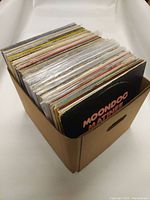 Box filled with over 30 vinyl records showing spines for identification, in cardboard box with handles on the sides.