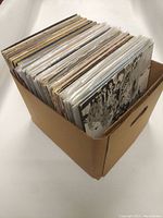 Cardboard box filled with multiple 12 inch vinyl records in protective sleeves.