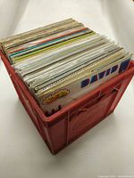 Red plastic crate filled with a collection of vinyl records showing colorful album covers along the top edges.