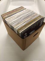Cardboard crate filled with approximately 40-50 loose vinyl records in protective plastic sleeves seen from above at an angle.