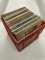 Red plastic crate filled with stacked 12" vinyl records, showing many album covers mostly obscured by back of crates.
