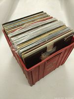 Red plastic crate filled with vinyl records standing upright, showing variety of album covers and spines