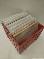 Red plastic crate filled with vinyl records packed in plastic sleeves, stacked upright.