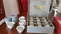 Photo showing boxed set of 16 fine porcelain sake cups with gold decorative pattern and additional unboxed sake cups with blue designs.