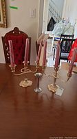 Two three-armed metal candle holders with pink taper candles, and one smaller single-stem metal candle holder on wooden table.