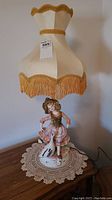 Full view of Capodimonte porcelain figurine lamp with cream fabric shade featuring gold trim and fringe, placed on a wooden table with lace doily.