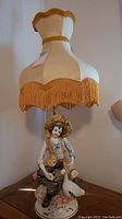 Full view of the porcelain table lamp showing the young lady figurine holding a duck beneath a cream-colored vintage lampshade with gold fringe.