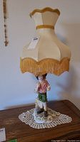 Full view of the Capodimonte porcelain lamp showing the young man figurine base and beige fringe-trimmed lampshade.