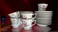 Set of 12 cups stacked with matching 12 saucers in background. Cups have white base with colorful floral designs and gold rims.