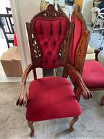 Photo showing two dining chairs close up, highlighting the elaborate carved wood frame and tufted red upholstery on the backrest and seat. One chair has arms, the other is armless.