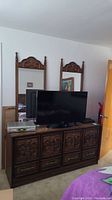 Front view of vintage dresser with television and other electronics on top, flanked by two tall matching carved wood mirrors. Shows detailed carving on drawer fronts.