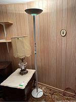 Full view of the white metal floor lamp standing next to a wooden table and another lamp in a room with wood panel wall and carpeted floor.