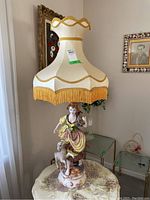 Full view of the tall hand painted ceramic lamp with figurine base and fabric lamp shade with gold trim and fringe.