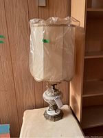 Full table lamp pictured sitting on a cream table against paneled wall, covered lamp shade in protective plastic, tag with number 604 attached