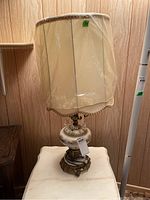 Full view of the lamp showing the beige fabric shade with protective plastic cover and the decorated glass and metal base.