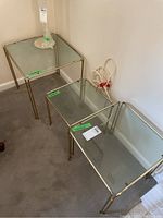 Set of three glass and brass nesting tables arranged side by side against wall on carpet. Largest table measures 19x19x19 as shown on tape label on glass surface. Visible slight smudges on glass and patina on brass frame.