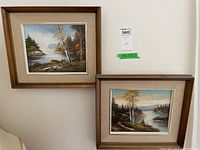 Two framed original wilderness scene paintings hanging on a white wall, showing their size and framing detail.