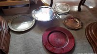 Five silver-plated serving items including two round trays with pierced edges and legs, a covered serving dish with Pyrex insert, a plain round tray, and a large red charger plate.