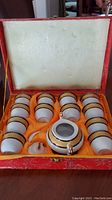 Open red decorative fabric box showing inside lined with satin orange and containing 12 small white ceramic tea cups, a teapot, and a spoon, all with matching black, gold, and orange decorative bands.