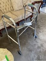 Full view of silver metal walker with grey handles and front wheels, placed on carpeted floor near sofa and wooden table.